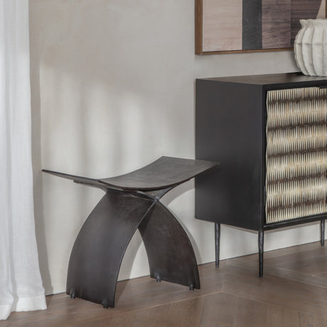 Libra Interiors Cast Aluminium Curved Stool in black with arched legs and concave seat, styled in a hallway beside a woven-front cabinet.