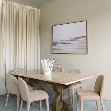 Libra Interiors Travertine Twin-Pedestal Dining Table in cream with etched antique brass legs, styled with six linen chairs beneath a coastal artwork.