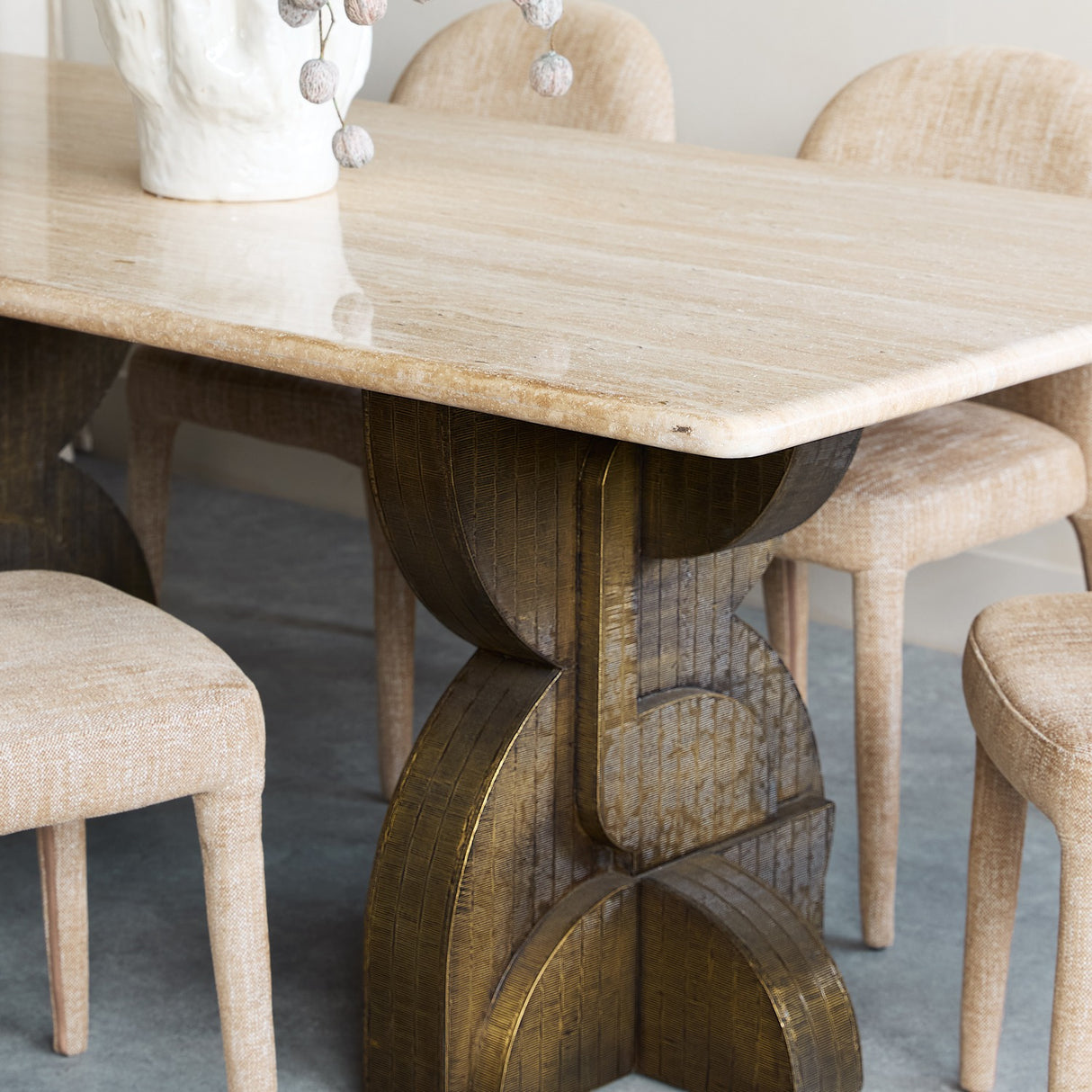 Libra Interiors Travertine Twin-Pedestal Dining Table in cream with a smooth stone top and sculptural etched antique brass pedestal, styled with linen chairs.