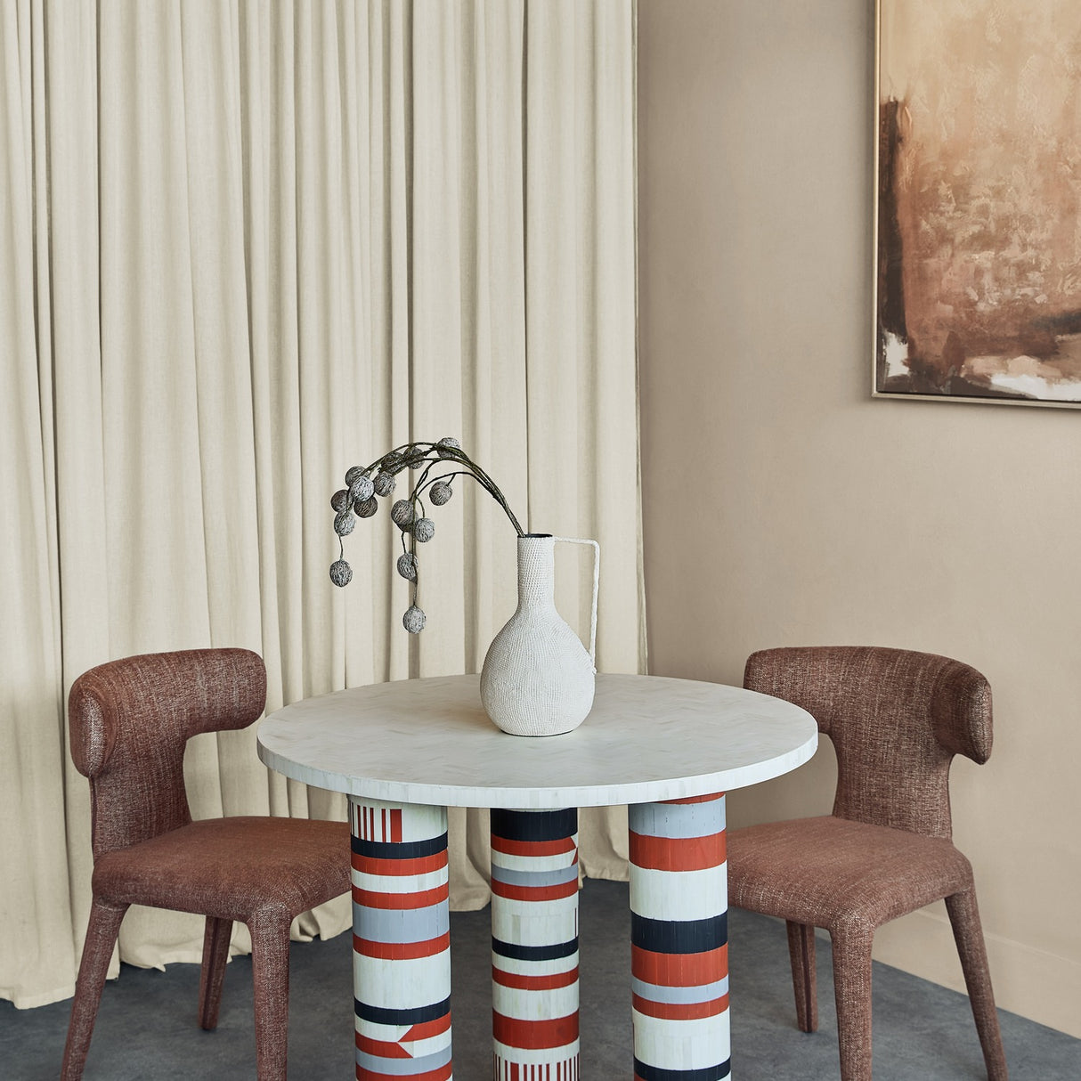 Libra Interiors Bone Inlay Round Dining Table with cream top and striped terracotta, black and grey legs, styled with a white vase between brown chairs.