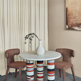 Libra Interiors Bone Inlay Round Dining Table with cream top and striped terracotta, black and grey legs, styled with a white vase between brown chairs.