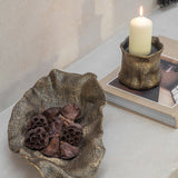 Libra Interiors woven-effect cast aluminium bowl in antique brass holding dried lotus pods, styled beside a matching candle holder on a book.