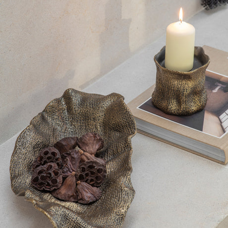 Libra Interiors woven-effect cast aluminium bowl in antique brass holding dried lotus pods, styled beside a matching candle holder on a book.