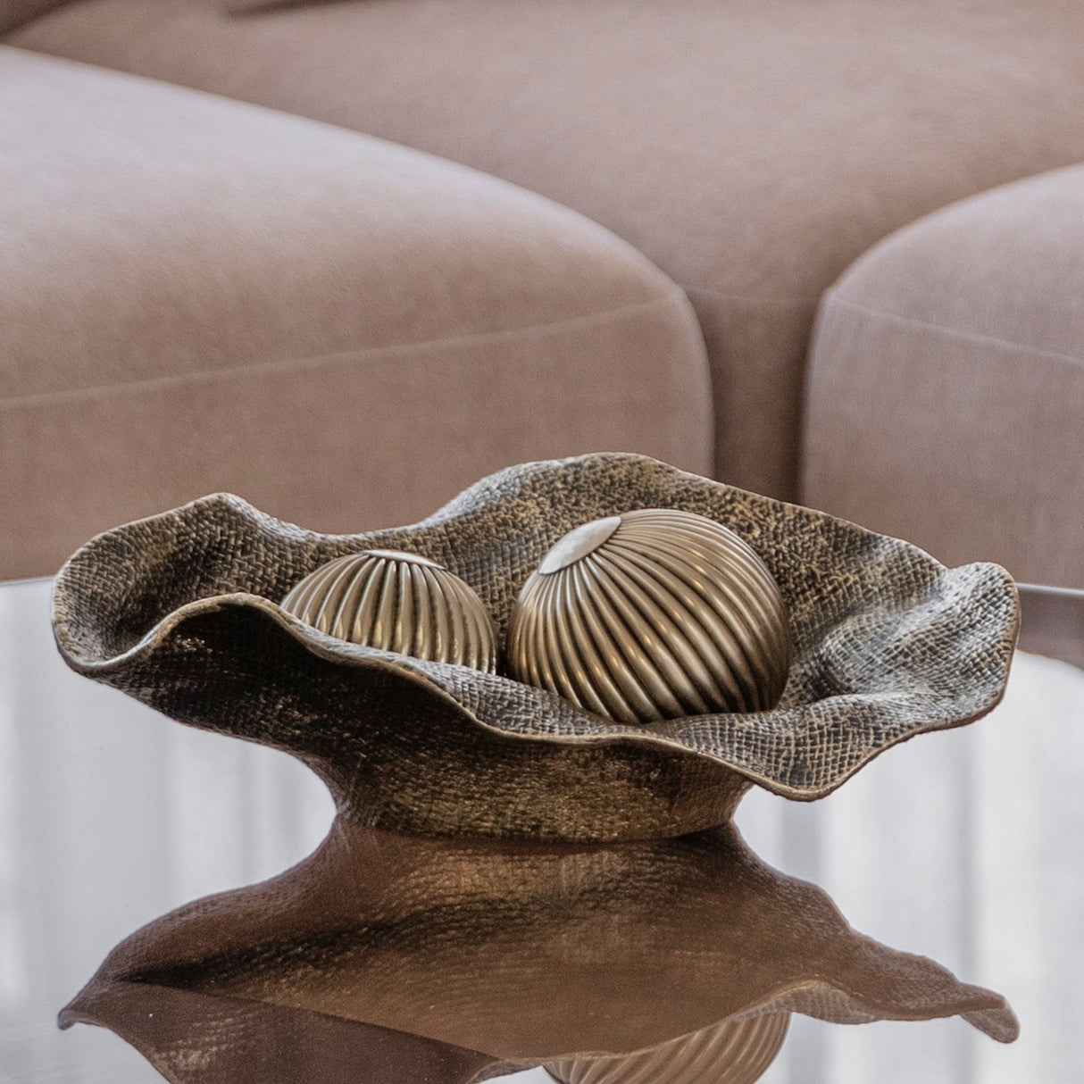 Libra Interiors woven-effect cast aluminium bowl in antique brass with rippled edges, holding two ribbed decorative spheres on a mirrored table by a beige sofa.
