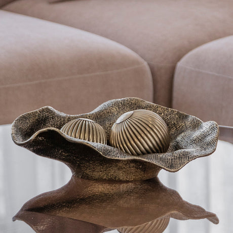 Libra Interiors woven-effect cast aluminium bowl in antique brass with rippled edges, holding two ribbed decorative spheres on a mirrored table by a beige sofa.
