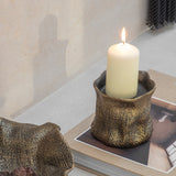 Libra Interiors woven-effect cast aluminium candle holder in antique brass, holding a lit ivory pillar candle atop a book in a styled setting.