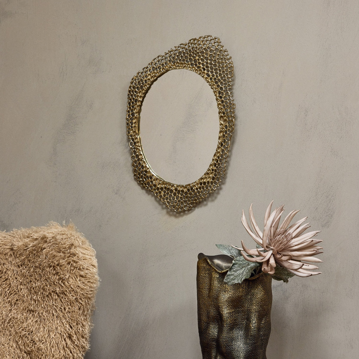 Libra Interiors Honeycomb cast aluminium wall mirror in gold with organic openwork frame, hung on a neutral wall above a bronze vase holding a pale bloom.