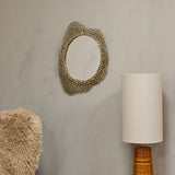 Libra Interiors Honeycomb cast aluminium wall mirror in gold with an irregular oval openwork frame, shown on a neutral wall beside a linen table lamp.