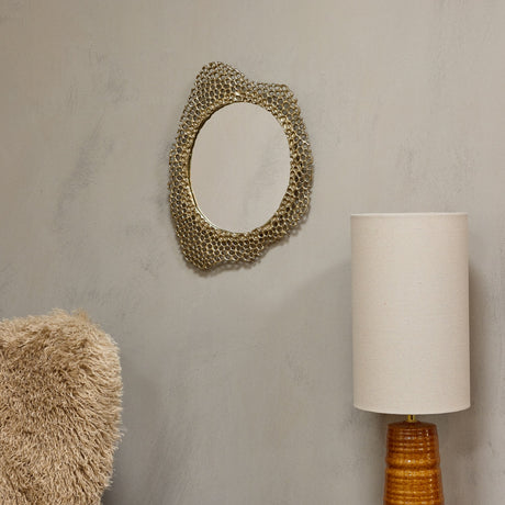 Libra Interiors Honeycomb cast aluminium wall mirror in gold with an irregular oval openwork frame, shown on a neutral wall beside a linen table lamp.