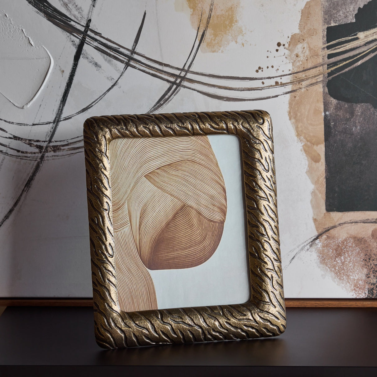 Libra Interiors Etched Cast Aluminium Photo Frame in antique brass with tiger-stripe texture, standing upright on a dark surface before abstract artwork.