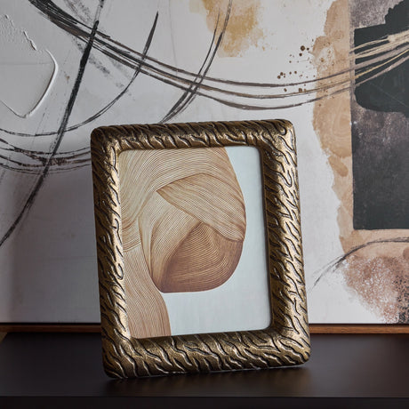Libra Interiors Etched Cast Aluminium Photo Frame in antique brass with tiger-stripe texture, standing upright on a dark surface before abstract artwork.