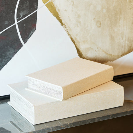 Libra Interiors beige linen book-style storage boxes stacked on a marble surface, set against abstract art in cream, gold and dark tones.