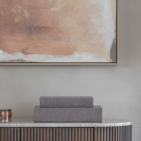 Libra Interiors Linen Weave book-style storage boxes in grey, stacked large-under-small on a fluted marble-topped console beneath a rust-toned abstract artwork.