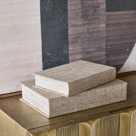 Libra Interiors Linen Weave book-shaped storage boxes in beige, stacked as a pair on a brass console beside a neutral abstract artwork.