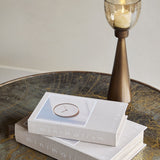 Libra Interiors Canvas Book-Style Storage Boxes in grey stacked on a bronzed round table beside a tall brass candle holder with amber glass.