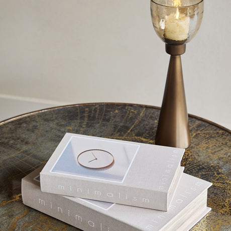Libra Interiors Canvas Book-Style Storage Boxes in grey stacked on a bronzed round table beside a tall brass candle holder with amber glass.
