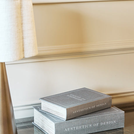 Libra Interiors Linen Book-Style Storage Boxes in grey stacked on a marble surface beside a lamp base, set against a cream panelled wall.