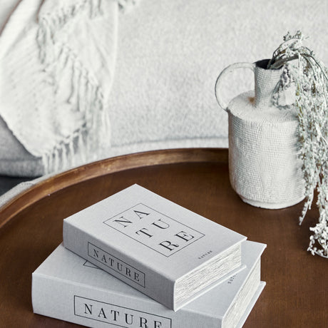 Libra Interiors Linen Book-Style Storage Boxes in white with black Nature typography, stacked on a wooden tray beside a textured vase of dried foliage.