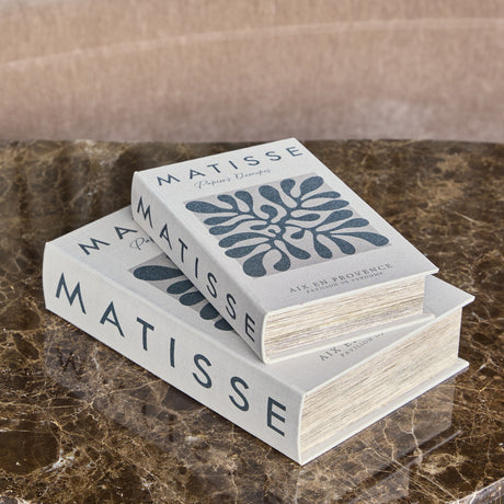 Libra Interiors Matisse book-style storage boxes in white linen with charcoal typography and paper-cutout motif, stacked on a brown marble surface.