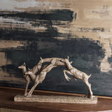 Libra Interiors Rustic Leaping Fawns resin sculpture in weathered brown, trio of arching deer on a rectangular base, set against a dark textured painting.
