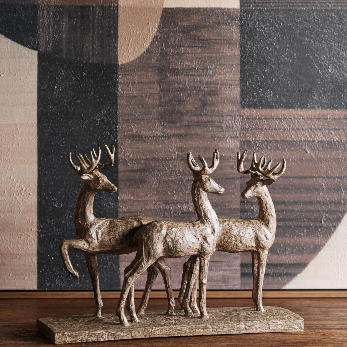 Libra Interiors Textured Trio of Stags Sculpture in brown resin with antlered figures on a shared base, styled on a wooden surface against abstract art.