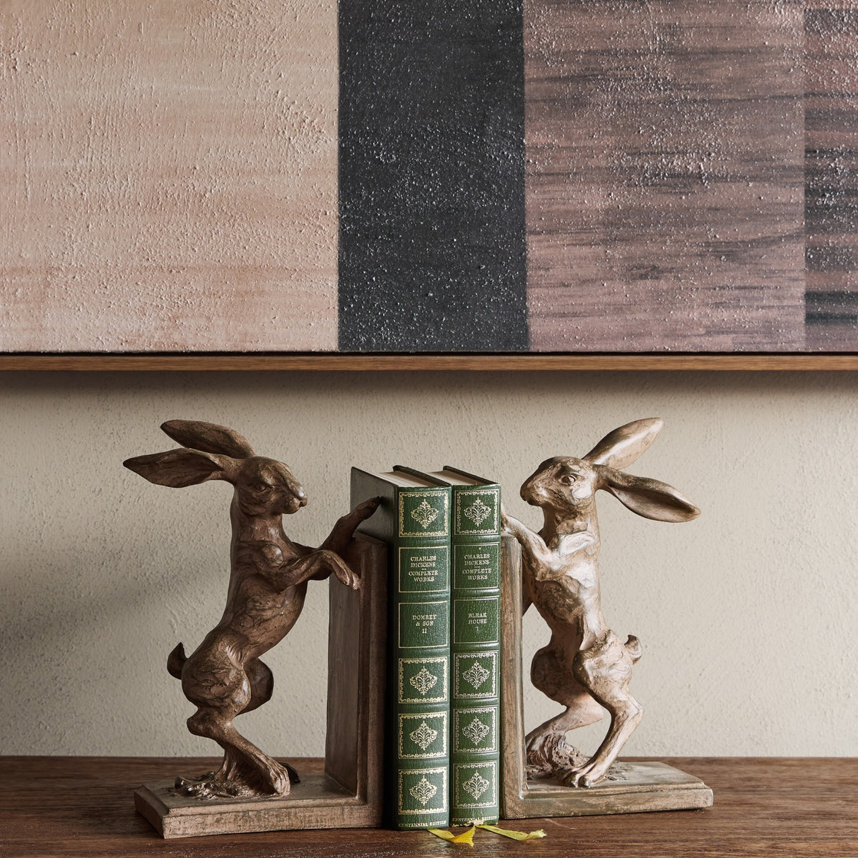 Libra Interiors Rustic Weathered Standing Hare Bookends in brown resin, framing green Charles Dickens volumes on a wooden shelf below abstract art.