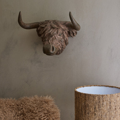 Libra Interiors Rustic Highland Cow resin wall art in brown, mounted on a taupe wall above a shaggy pouffe and woven basket lamp.