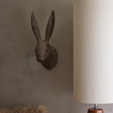 Libra Interiors Rustic Weathered Hare Head wall art in brown resin, mounted on a plaster wall beside a cream cylindrical table lamp in a hallway setting.