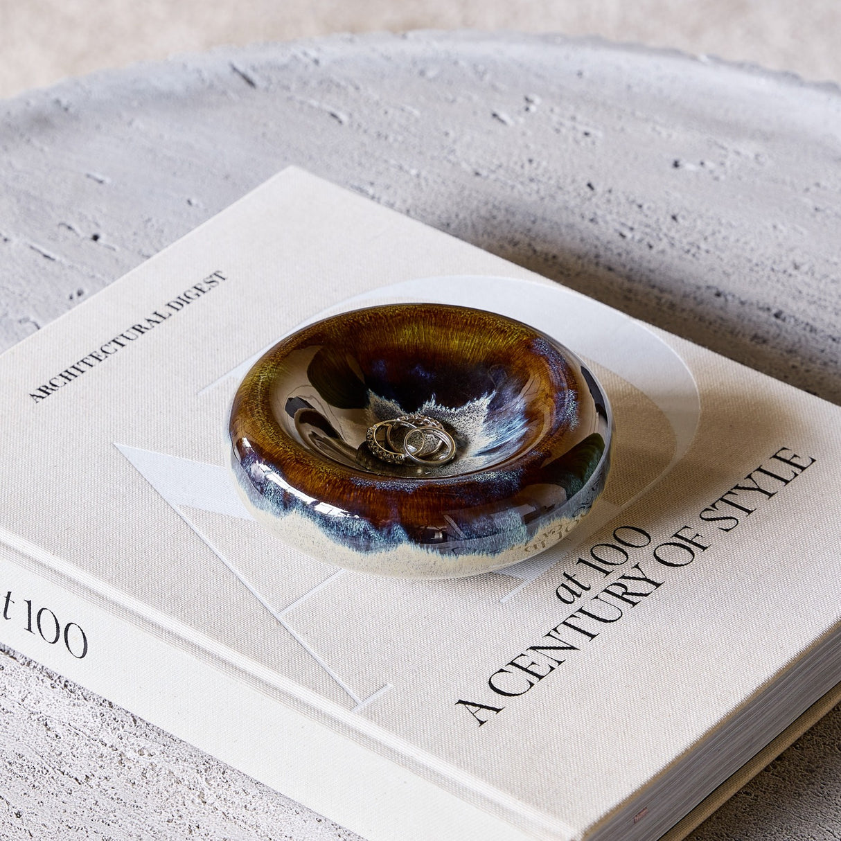 Libra Interiors Reactive Glaze Stoneware Bowl in blue and smoky grey, holding rings, styled on an Architectural Digest book beside a stone tray.