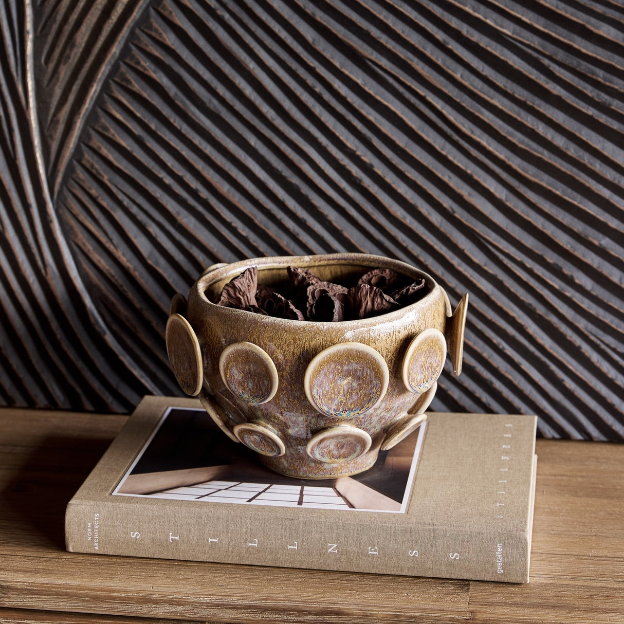 Libra Interiors Reactive Glaze Raised Circle stoneware planter in mottled brown, holding dried bark, displayed on a linen book against a carved wood panel.