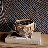 Libra Interiors Reactive Glaze Raised Circle stoneware planter in mottled brown, holding dried bark, displayed on a linen book against a carved wood panel.