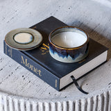 Libra Interiors Reactive Glaze Ceramic Rose Candle in blue with lid beside it, resting on a Monet book atop a textured pale stone table.