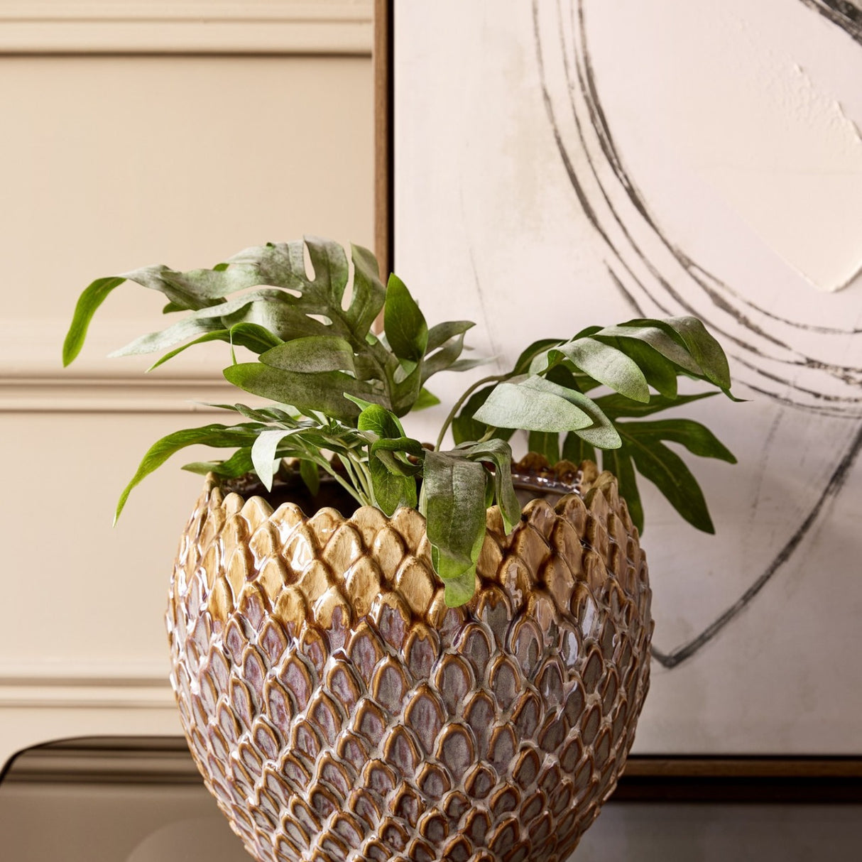 Libra Interiors Scale-Textured Ceramic Planter in brown, holding leafy foliage on a console beside abstract framed art and cream panelling.
