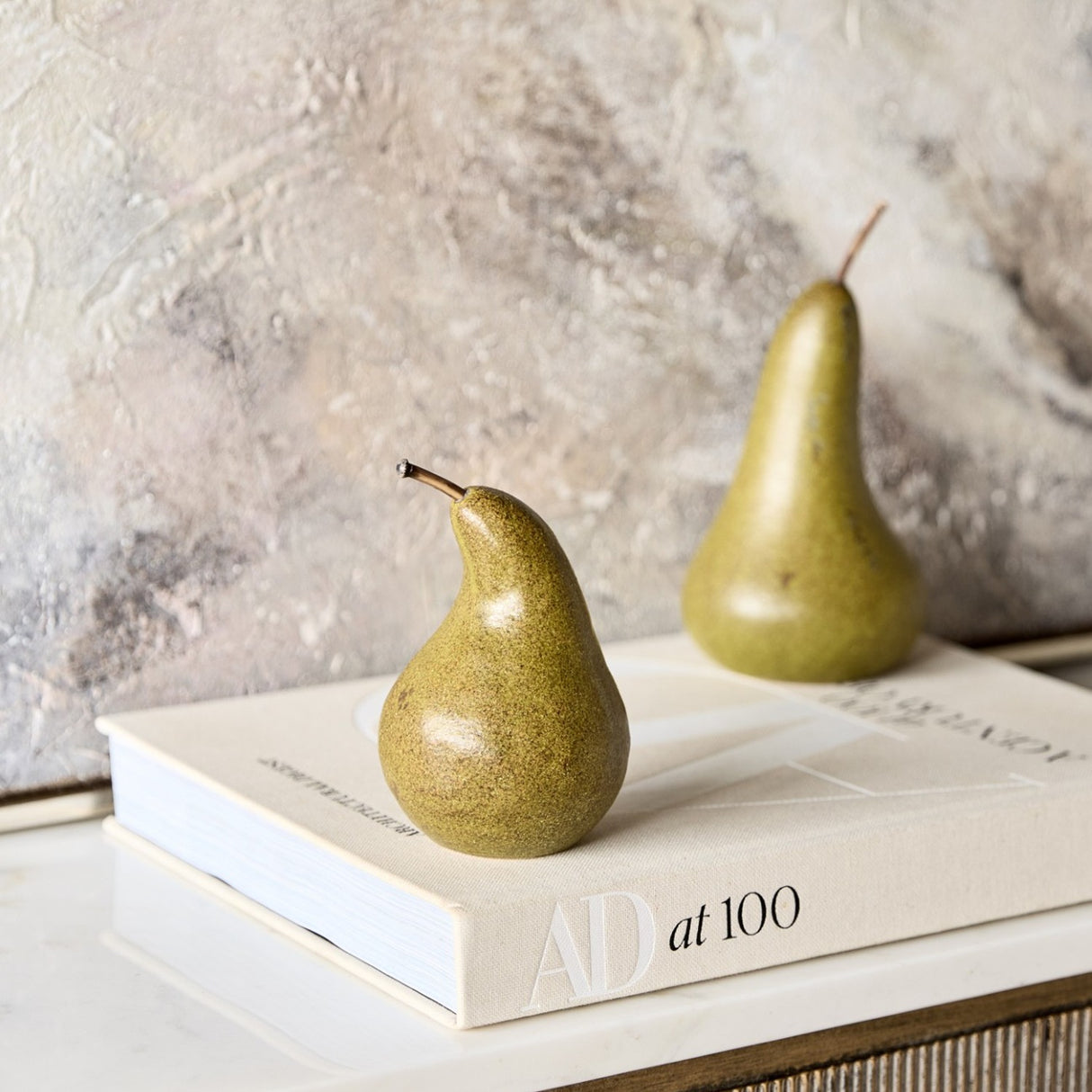 Libra Interiors Speckled Ceramic Pear Sculptures in green and brown, displayed on an AD at 100 book against a textured plaster wall.