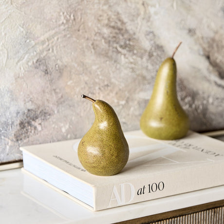 Libra Interiors Speckled Ceramic Pear Sculptures in green and brown, displayed on an AD at 100 book against a textured plaster wall.
