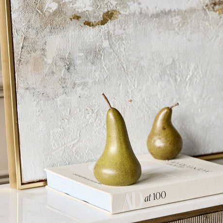 Two Libra Interiors speckled green ceramic pear sculptures with slim stems, displayed on an AD at 100 book beside a textured gold and grey artwork.