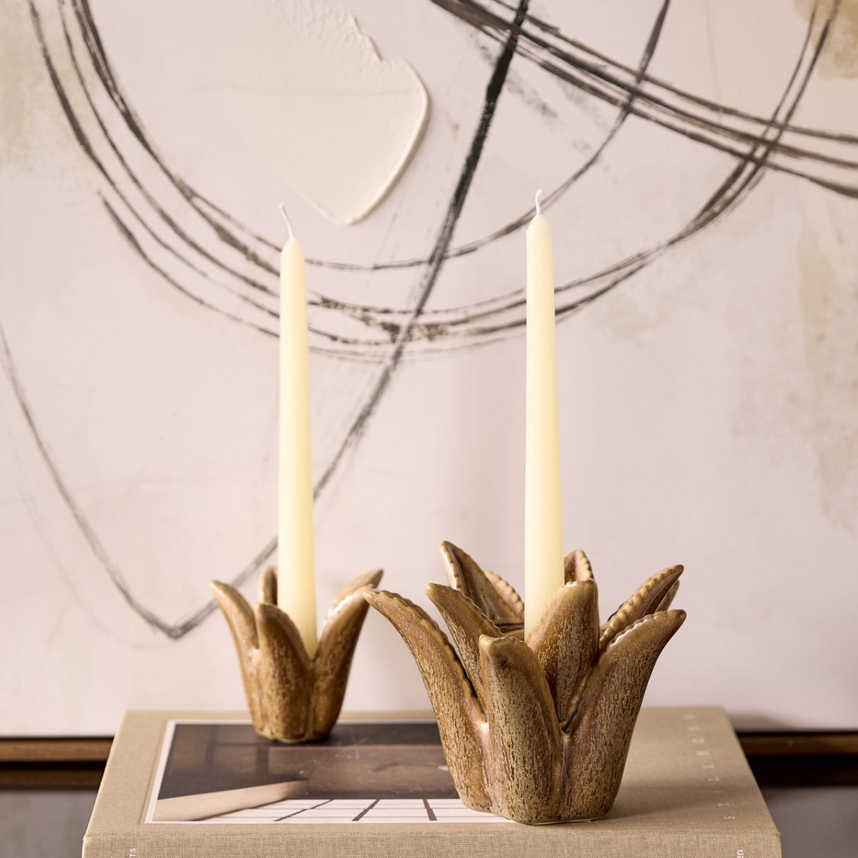 Libra Interiors Petal reactive glaze ceramic candle holders in brown, sculpted as tropical blooms holding cream taper candles atop a book by abstract art.