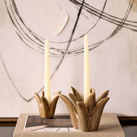 Libra Interiors Petal reactive glaze ceramic candle holders in brown, sculpted as tropical blooms holding cream taper candles atop a book by abstract art.