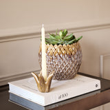 Libra Interiors Petal reactive glaze ceramic candle holder in brown with a tapered candle, displayed on stacked books beside a pineapple-shaped planter.