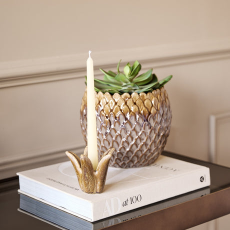 Libra Interiors Petal reactive glaze ceramic candle holder in brown with a tapered candle, displayed on stacked books beside a pineapple-shaped planter.