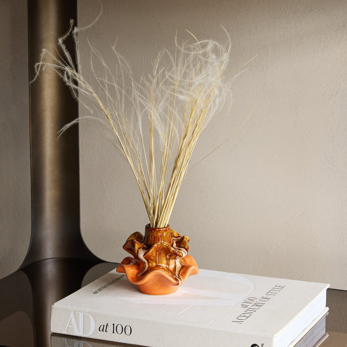 Libra Interiors Reactive Glaze Ceramic Wavy Small Vase in brown and orange with dried grasses, styled on a design book beside a bronze frame.