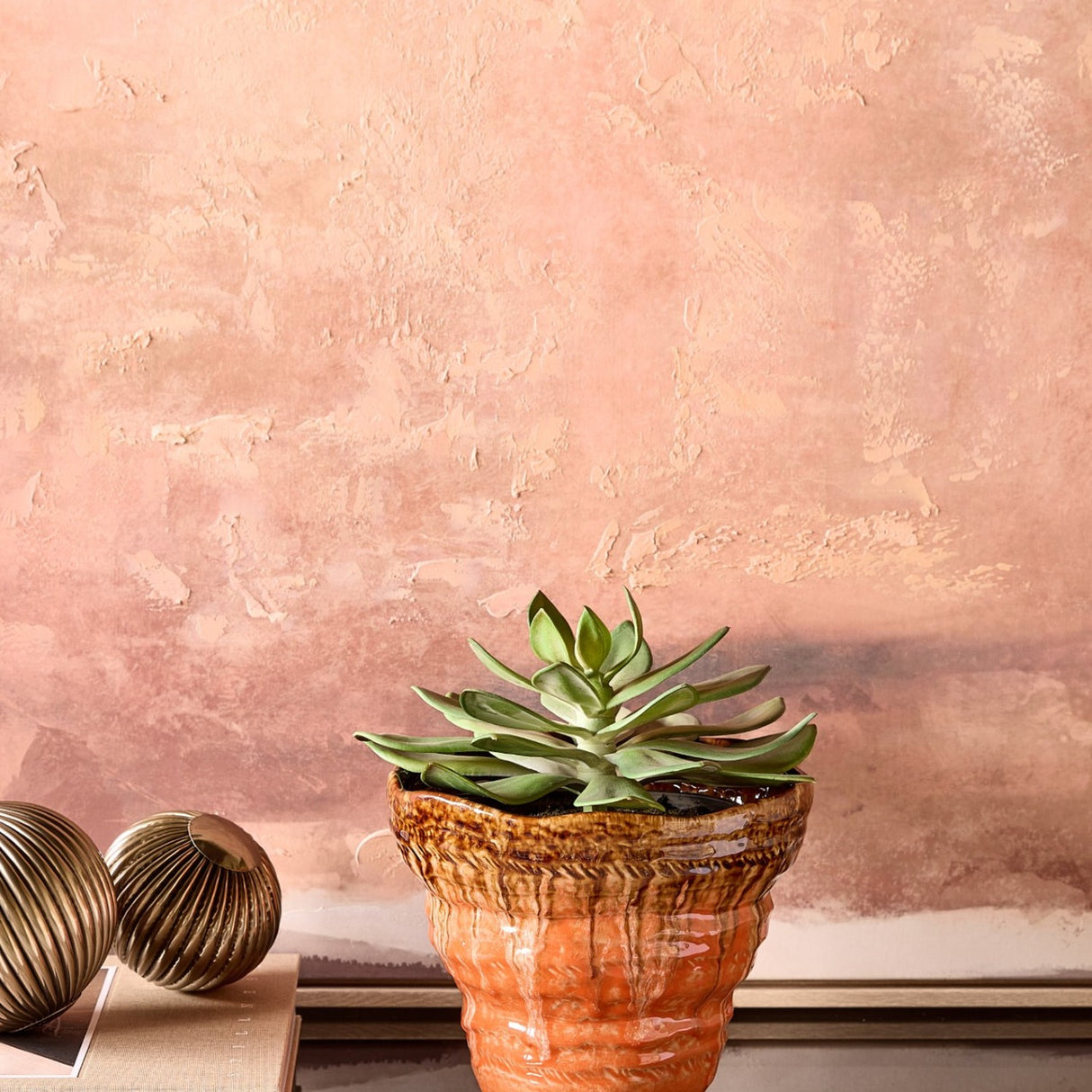 Libra Interiors Reactive Glaze Ceramic Planter in brown and orange holds a succulent beside brass ornaments against a textured pink plaster wall.