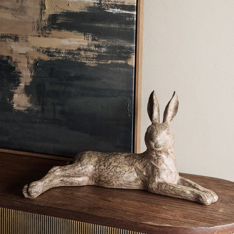 Libra Interiors Rustic Lying Hare resin sculpture in weathered brown, ears upright, displayed on a wooden console beside a dark textured artwork.