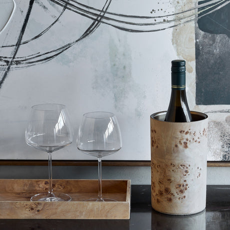 Libra Interiors Burl Veneer Brass-Lined Wine Rack in natural finish holding a wine bottle beside two stemmed glasses on a wooden tray with abstract art behind.