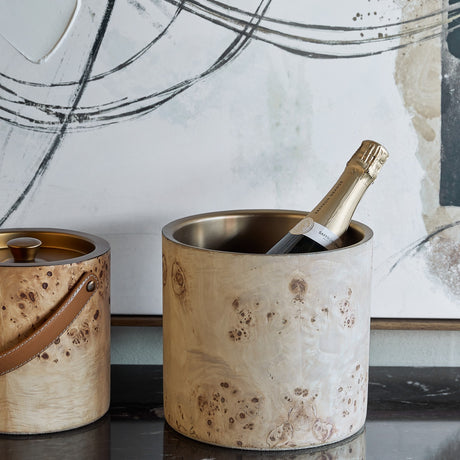 Libra Interiors Burl Veneer Brass-Lined Wine Rack in natural finish holding a champagne bottle, shown beside a matching ice bucket on a dark surface.