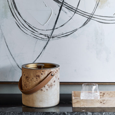 Libra Interiors Burlwood Veneer Brass Lidded Ice Bucket with tan leather handle, styled on a dark surface beside a tumbler and abstract artwork.