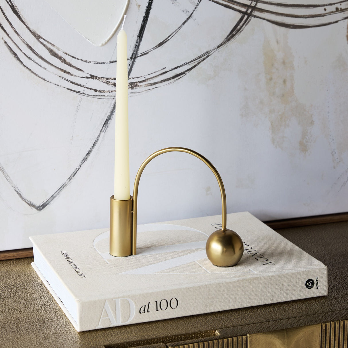 Libra Interiors Arch Gold Taper Candle Holder in polished iron with curved arch and spherical counterweight, holding an ivory taper on a book beside artwork.