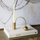 Libra Interiors Arch Gold Taper Candle Holder in polished iron with curved arch and spherical counterweight, holding an ivory taper on a book beside artwork.