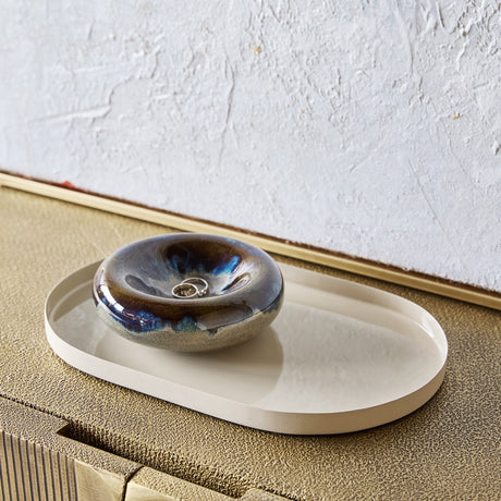 Libra Interiors Enamelled Oval Iron Serving Tray in cream, holding a glossy blue ceramic ring dish with a silver ring, on a textured gold console.