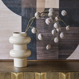 Libra Interiors Tiered Enamel Iron Vase in cream with stacked rounded tiers, holding a dried berry branch on a brass sideboard against abstract art.